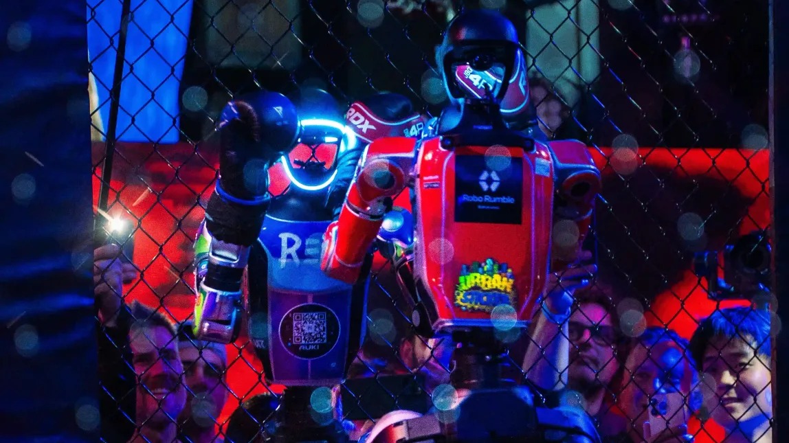 A sponsor-covered humanoid robot in the cage at a REK event, crowd pressed up against the wire